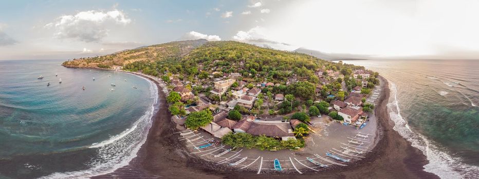 Aerial view of Amed beach in Bali, Indonesia. Traditional fishing boats called jukung on the black sand beach and Mount Agung volcano in the background, partially covered by clouds.