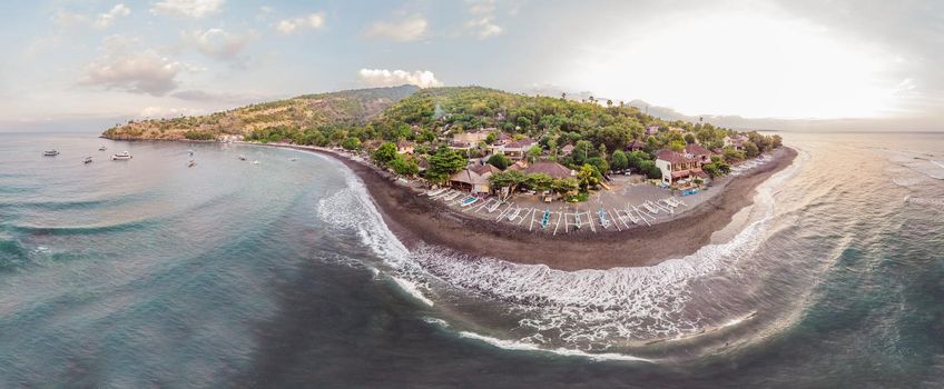 Aerial view of Amed beach in Bali, Indonesia. Traditional fishing boats called jukung on the black sand beach and Mount Agung volcano in the background, partially covered by clouds.