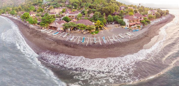 Aerial view of Amed beach in Bali, Indonesia. Traditional fishing boats called jukung on the black sand beach and Mount Agung volcano in the background, partially covered by clouds.