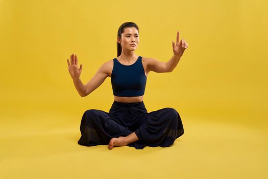 Front view of sporty girl sitting with crossed legs on floor, raising hands, holding finger up, looking forward. Young female practicing yoga pose. Isolated on yellow background.