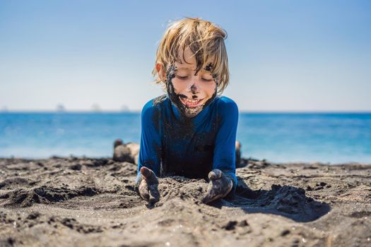 Black Friday concept. Smiling boy with dirty Black face sitting and playing on black sand sea beach before swimming in ocean. Family active lifestyle, and water leisure on summer vacation with kids. Black Friday, sales of tours and airline tickets or goods.