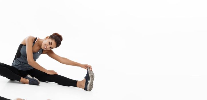 Sport, training, lifestyle and Fitness concept - portrait of beautiful happy African American woman stretching leg while sitting. Isolated on white studio background