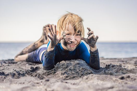 Black Friday concept. Smiling boy with dirty Black face sitting and playing on black sand sea beach before swimming in ocean. Family active lifestyle, and water leisure on summer vacation with kids. Black Friday, sales of tours and airline tickets or goods.