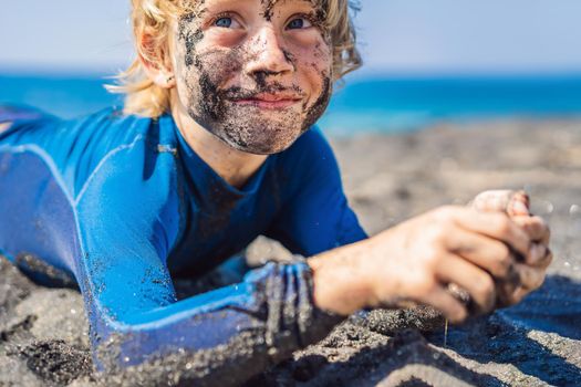 Black Friday concept. Smiling boy with dirty Black face sitting and playing on black sand sea beach before swimming in ocean. Family active lifestyle, and water leisure on summer vacation with kids. Black Friday, sales of tours and airline tickets or goods.