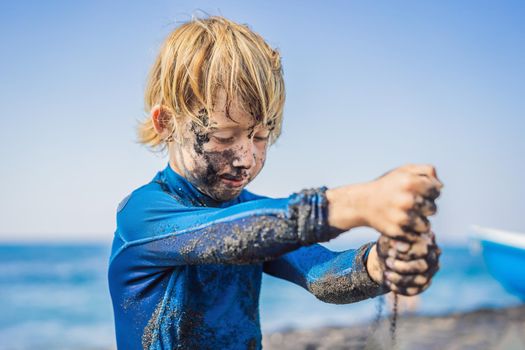 Black Friday concept. Smiling boy with dirty Black face sitting and playing on black sand sea beach before swimming in ocean. Family active lifestyle, and water leisure on summer vacation with kids. Black Friday, sales of tours and airline tickets or goods.