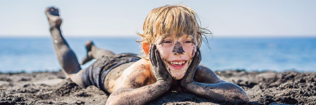 BANNER, LONG FORMAT Black Friday concept. Smiling boy with dirty Black face sitting and playing on black sand sea beach before swimming in ocean. Family active lifestyle, and water leisure on summer vacation with kids. Black Friday, sales of tours and airline tickets or goods.