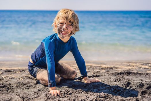 Black Friday concept. Smiling boy with dirty Black face sitting and playing on black sand sea beach before swimming in ocean. Family active lifestyle, and water leisure on summer vacation with kids. Black Friday, sales of tours and airline tickets or goods.