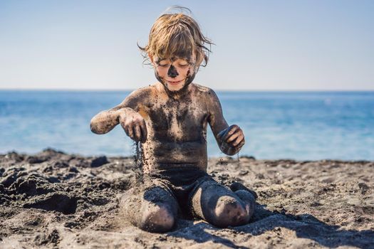 Black Friday concept. Smiling boy with dirty Black face sitting and playing on black sand sea beach before swimming in ocean. Family active lifestyle, and water leisure on summer vacation with kids. Black Friday, sales of tours and airline tickets or goods.