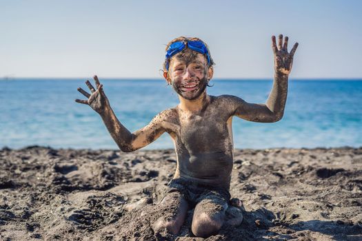 Black Friday concept. Smiling boy with dirty Black face sitting and playing on black sand sea beach before swimming in ocean. Family active lifestyle, and water leisure on summer vacation with kids. Black Friday, sales of tours and airline tickets or goods.
