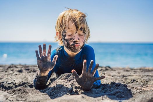 Black Friday concept. Smiling boy with dirty Black face sitting and playing on black sand sea beach before swimming in ocean. Family active lifestyle, and water leisure on summer vacation with kids. Black Friday, sales of tours and airline tickets or goods.