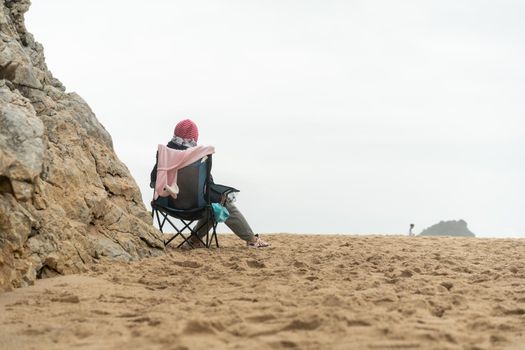 Thoughtful elderly woman sitting touristic chair on sea beach looking horizon Loneliness and old age concept Autumn of life Cold weather warm clothes