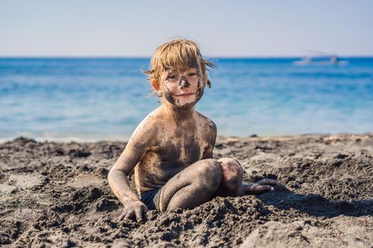 Black Friday concept. Smiling boy with dirty Black face sitting and playing on black sand sea beach before swimming in ocean. Family active lifestyle, and water leisure on summer vacation with kids. Black Friday, sales of tours and airline tickets or goods.