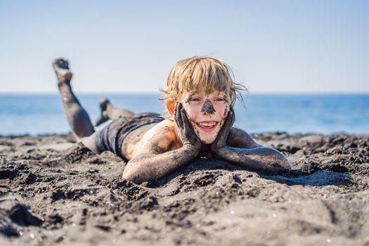 Black Friday concept. Smiling boy with dirty Black face sitting and playing on black sand sea beach before swimming in ocean. Family active lifestyle, and water leisure on summer vacation with kids. Black Friday, sales of tours and airline tickets or goods.