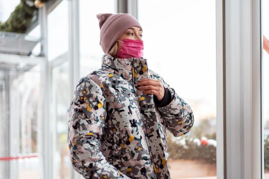 European woman in pink medical mask dressed winter clothes quarantine, coronavirus pandemic, shopping, shop, street. wearing winter coat looking through window on city street