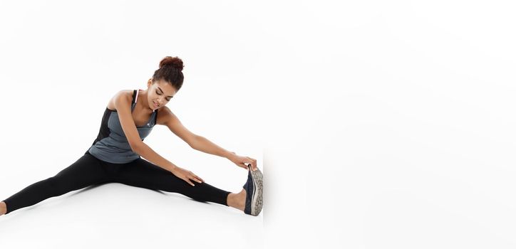 Sport, training, lifestyle and Fitness concept - portrait of beautiful happy African American woman stretching leg while sitting. Isolated on white studio background