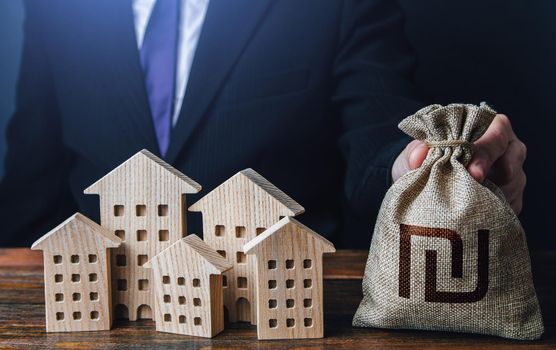 A businessman puts a israeli shekel money bag near the houses. Construction industry, rental business and hotel tourism. Municipal budget of the city. Official. Investments in real estate assets.