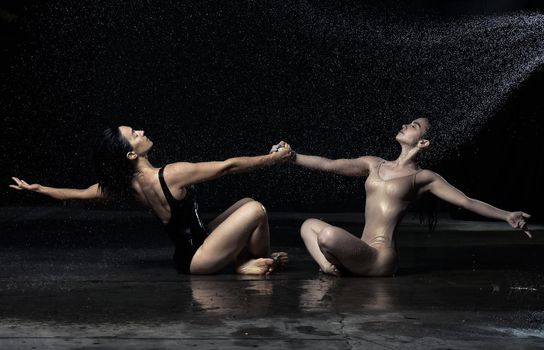 two beautiful women of Caucasian appearance with dark hair are sitting on the asphalt in drops of water on a black background, holding hands and looking up. People are wearing bodysuits