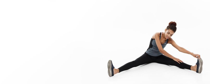 Sport, training, lifestyle and Fitness concept - portrait of beautiful happy African American woman stretching leg while sitting. Isolated on white studio background