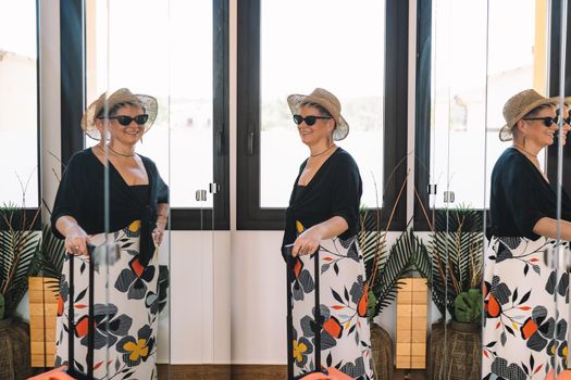 mature woman traveller, looking in the mirror while trying on her sunglasses for a walk along the coast on her summer holiday. bedroom, natural light, dressing room with mirrors.