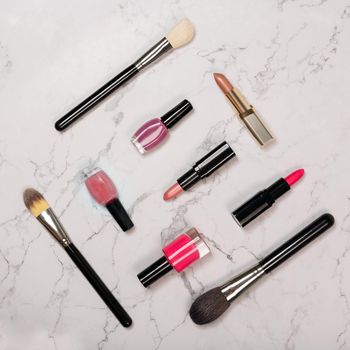 Flat lay of cosmetic products including lipsticks, nail varnish, makeup brushes on white marble background