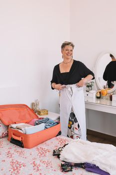 mature woman packing her luggage for summer holiday. business woman travelling. trip and hobby concept. looking at camera.. Room, illuminated by natural light, suitcase with clothes. vertical.