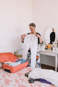 smiling mature woman preparing her luggage for a family trip in the summer. mother packing her children's suitcase. concept of leisure and hobby. Vertical, Room, illuminated by natural light, suitcase with clothes.