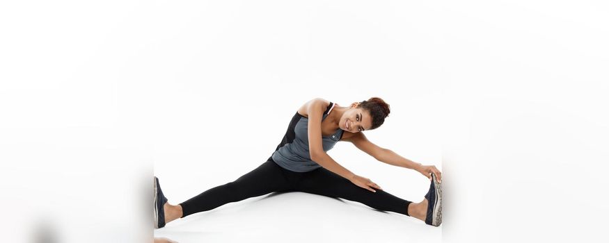 Sport, training, lifestyle and Fitness concept - portrait of beautiful happy African American woman stretching leg while sitting. Isolated on white studio background