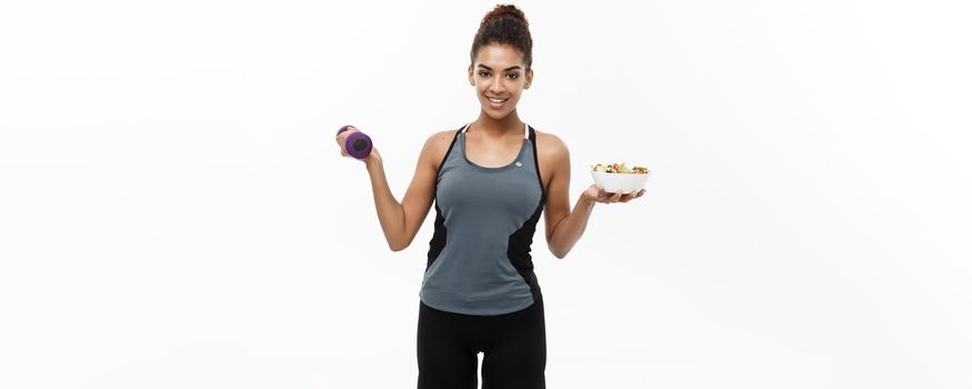 Healthy and Fitness concept - Beautiful sporty African American on diet holding dumbbell and fresh salad on hands. Isolated on white studio background