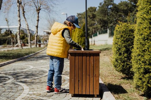 Schoolboy kid throwing the trash into dumpster. Boy using recycling bin to throw away the litter. Caucasian child recycles the junk into the trash-can..