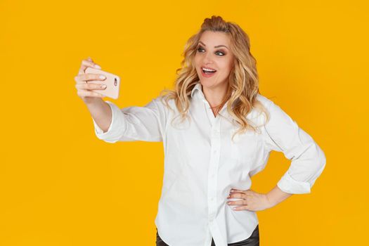 A blond woman in a white middle-aged shirt takes a selfie on a smartphone, holds a mobile phone in her outstretched hand and tries to look good in a photo on a yellow background.