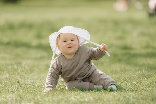 A 7-month child in the white panama hat is having fun in the meadow. An infant girl is crawling on the grass.