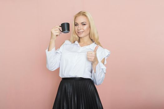 Beauty, fashion portrait. Elegant business style. Portrait of a beautiful blonde woman in white blouse and black skirt posing at studio on a pink background. Holding black cup in her hands. Drinking coffee, tea
