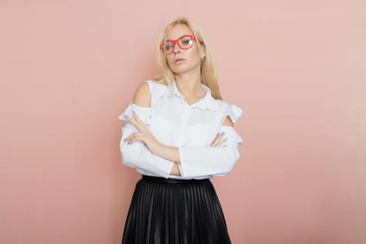 Beauty, fashion portrait. Elegant business style. Portrait of a beautiful blonde woman in white blouse and black skirt posing at studio on a pink background. Wearing red glasses