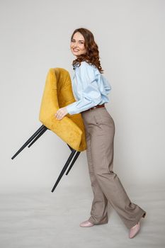 A girl in a blue shirt and trousers holds a yellow chair in her hands.