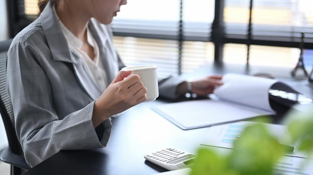 Cropped shot female manager drinking coffee and reading reports at office desk.