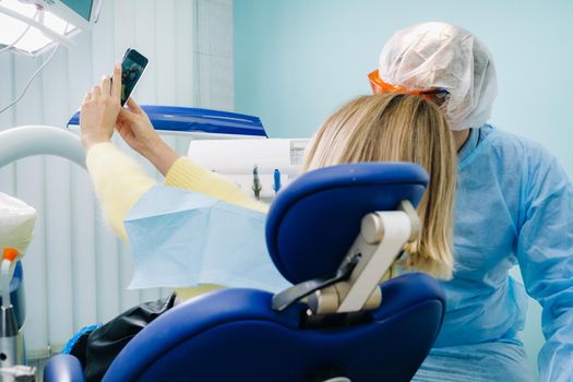 a dentist in a protective mask sits next to a patient and takes a selfie photo while working.