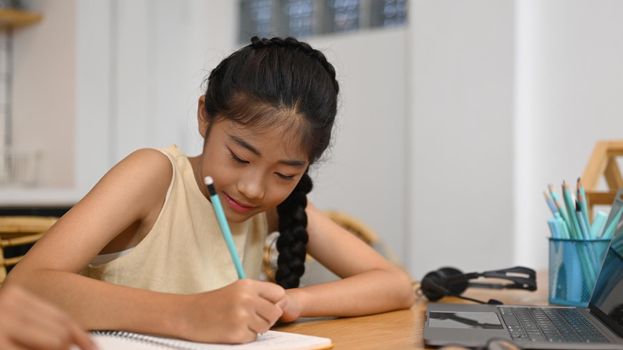 Asian school girl distance learning online, doing homework while sitting at kitchen table.