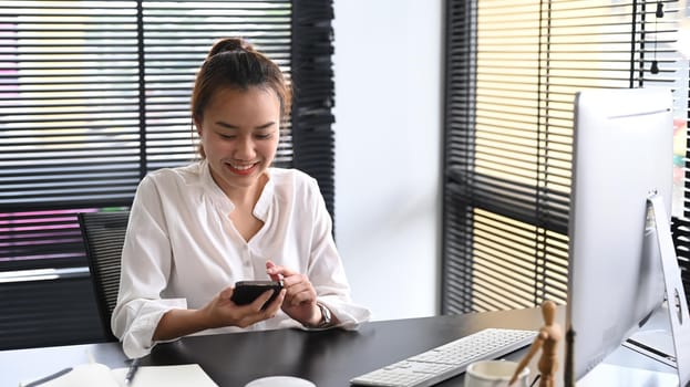 Happy millennial asian woman sitting at her workplace and using mobile phone for communicating in social network.