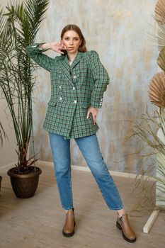 A girl in a green fashionable jacket and blue jeans on a studio background with plants.