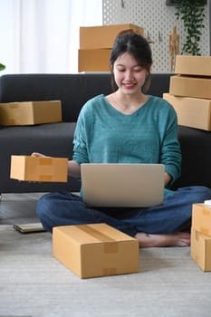 Smiling young man checking product purchase order on laptop computer and preparing parcel for delivery.