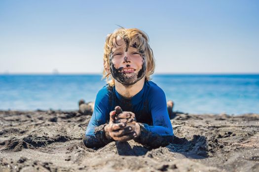 Black Friday concept. Smiling boy with dirty Black face sitting and playing on black sand sea beach before swimming in ocean. Family active lifestyle, and water leisure on summer vacation with kids. Black Friday, sales of tours and airline tickets or goods.