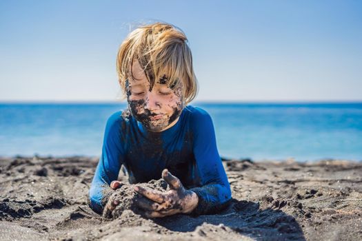 Black Friday concept. Smiling boy with dirty Black face sitting and playing on black sand sea beach before swimming in ocean. Family active lifestyle, and water leisure on summer vacation with kids. Black Friday, sales of tours and airline tickets or goods.