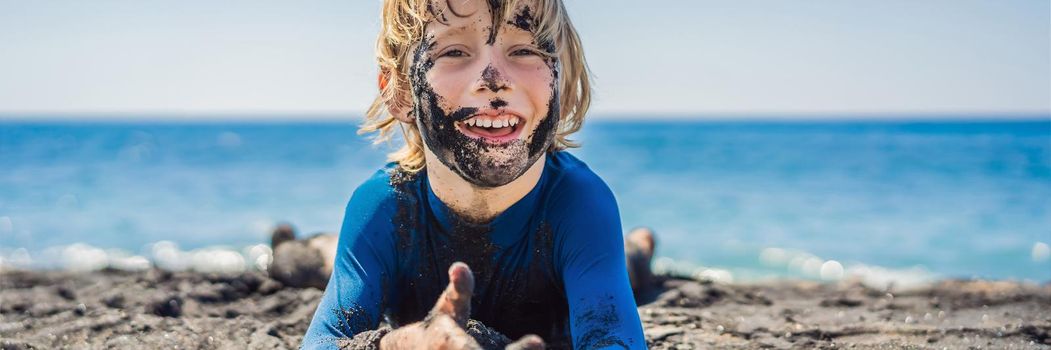 BANNER, LONG FORMAT Black Friday concept. Smiling boy with dirty Black face sitting and playing on black sand sea beach before swimming in ocean. Family active lifestyle, and water leisure on summer vacation with kids. Black Friday, sales of tours and airline tickets or goods.