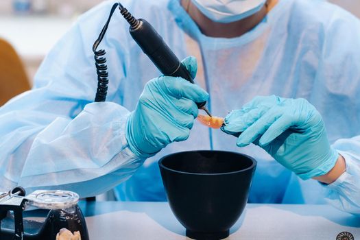 A masked and gloved dental technician works on a prosthetic tooth in his lab.