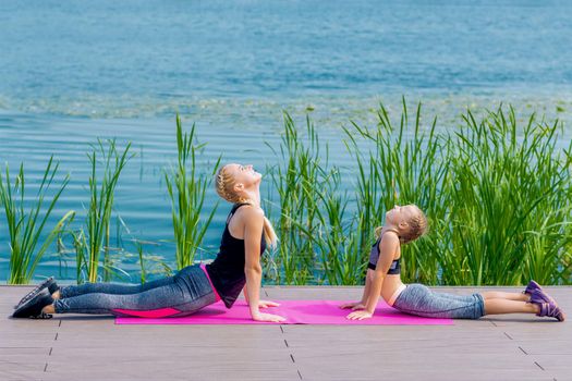 Little girt and woman are doing exercises on the grass at the shore of the lake.