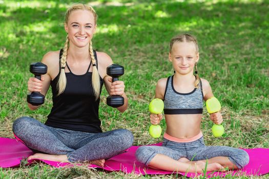 Young mother with her little daughter are training with apple dumbbells in the park.