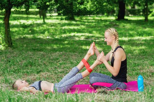 Mother is training legs of daughter by fitness gum on mat in the park.