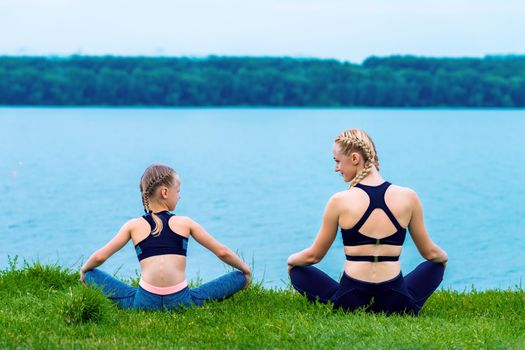 Mother and daughter doing yoga exercises on grass of the shore of the lake.