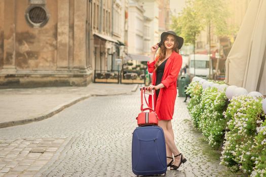 Cheerful white woman standing on the old street wearing hat with suitcase smiling at camera on background of city.