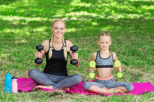 Young mother with her little daughter are training with apple dumbbells in the park.